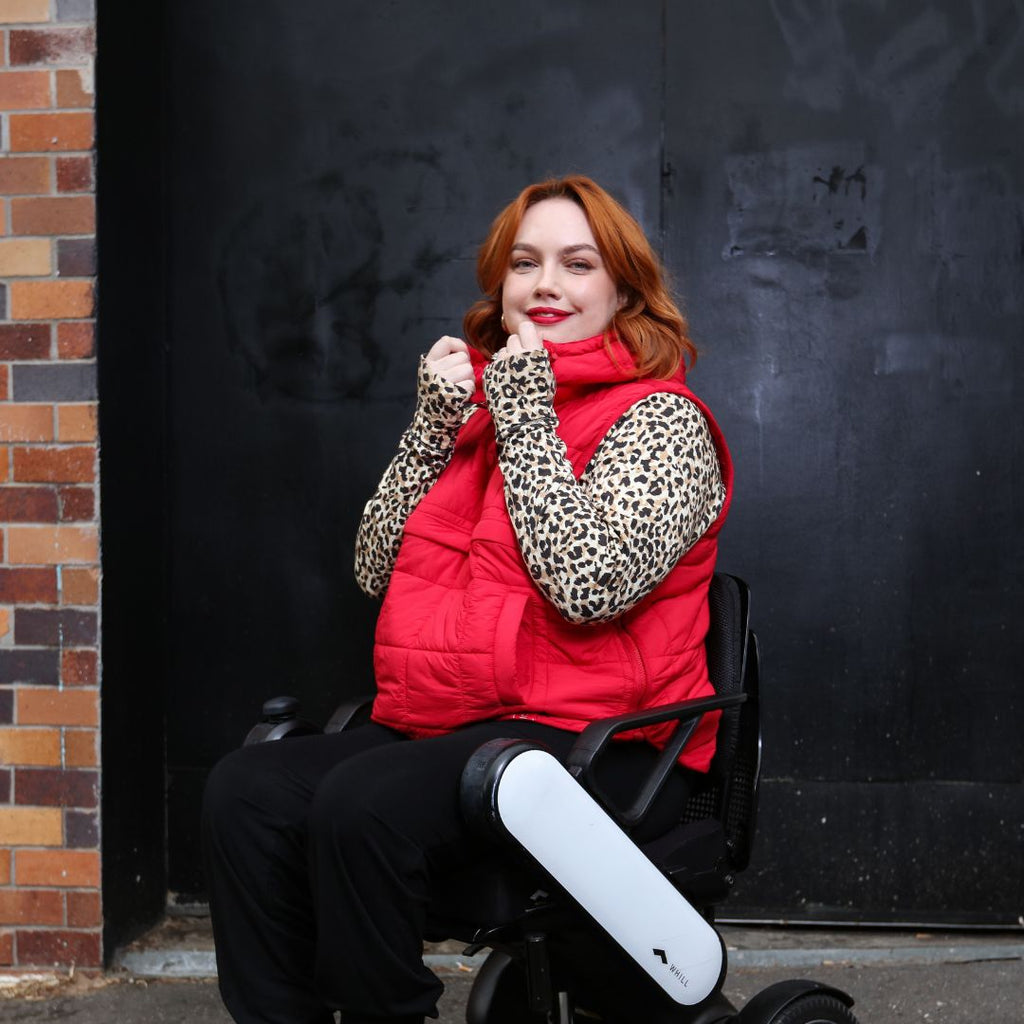 Female with red hair in a wheelchair wearing a leopard print long sleeve top, bright red puffer vest and black bottoms. Christina Stephens Adaptive Clothing Australia. 