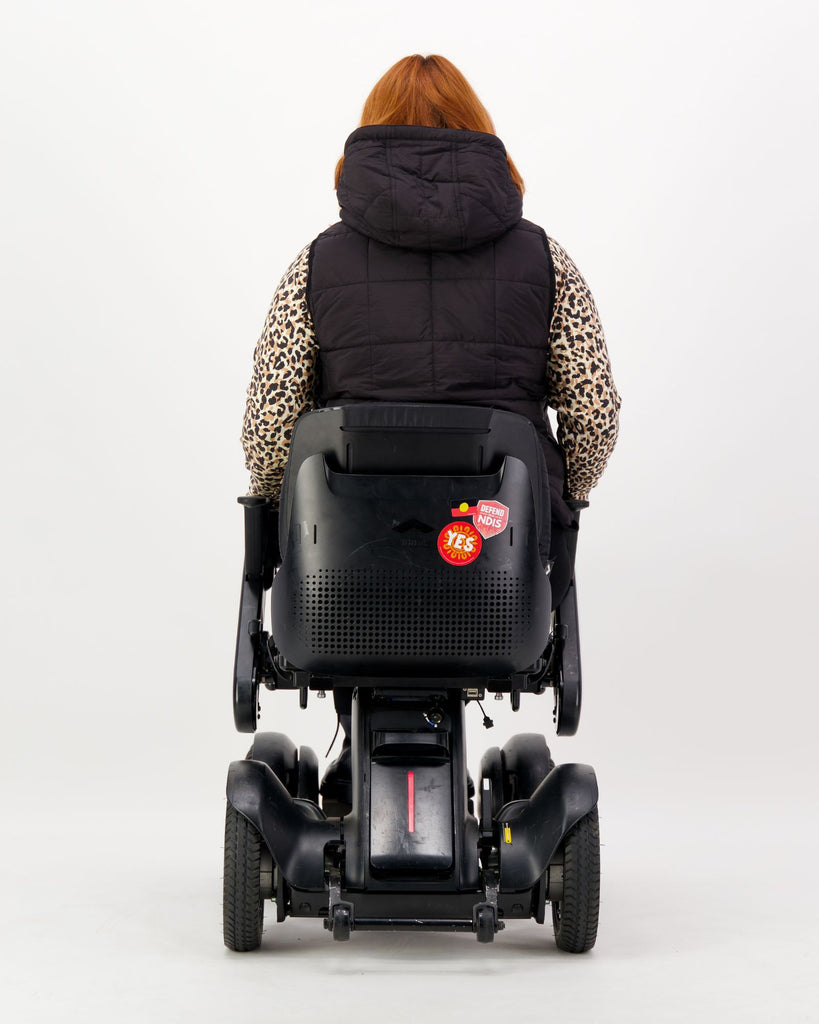 Seated female wearing a black puffer vest over black clothing while seated in a power wheelchair. Christina Stephens Adaptive Clothing Australia.