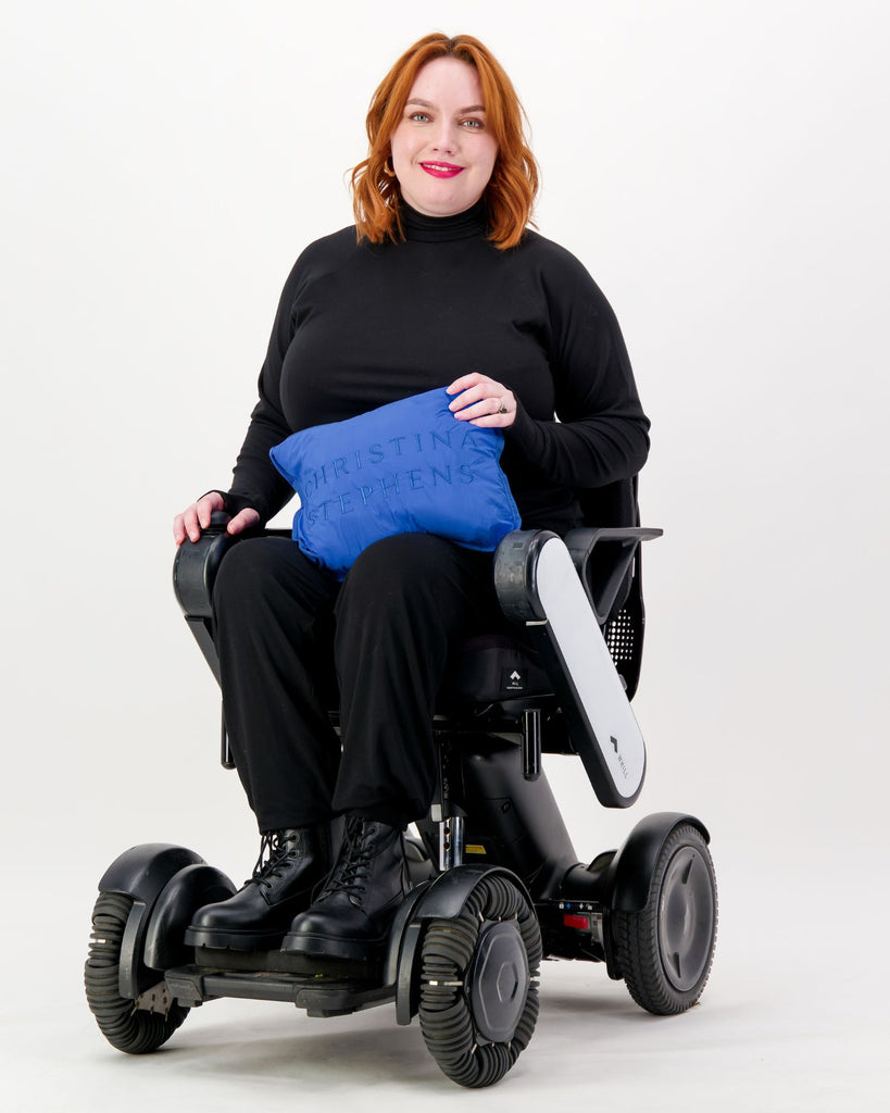 Seated female wearing a black thermal top over black clothing while seated in a power wheelchair. She is holding a pocket purse packed with the puffer vest in cobolt blue. Christina Stephens Adaptive Clothing Australia.