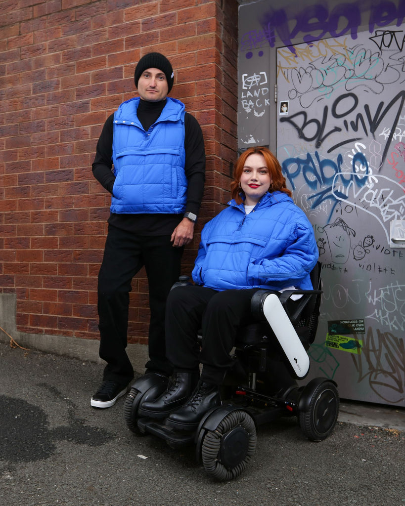 Standing male and seated female wearing cobalt blue puffer vest and jacket over black clothing. Christina Stephens Adaptive Clothing Australia.