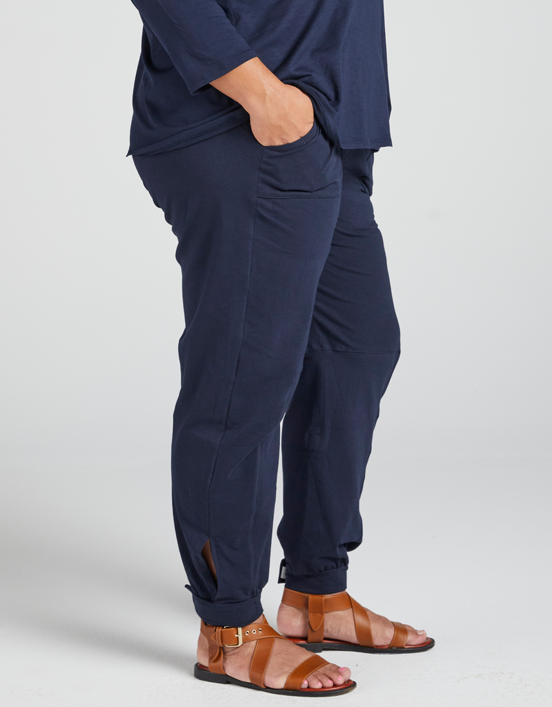 Model wearing navy blue pants and brown sandals. Pants are tapered to the ankle, and display square pocket details. Model's right hand is in her pocket. Standing. Side facing. 