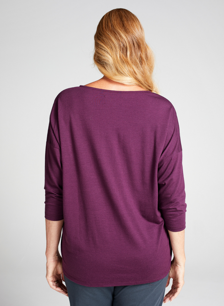 Blonde woman wearing a grape (purple) round neck, loose top with 3/4 length sleeves. Back facing. 