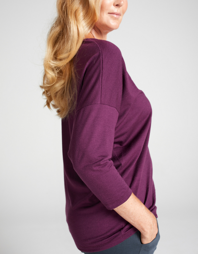 Blonde woman wearing a grape (purple) round neck, loose top with 3/4 length sleeves. Standing. Side facing. 