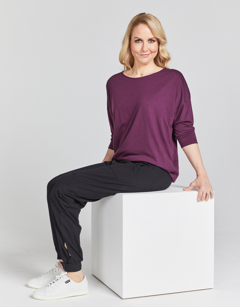 Seated blonde woman  wearing a grape (purple) round neck, loose top with 3/4 length sleeves and black pants. Side facing. 