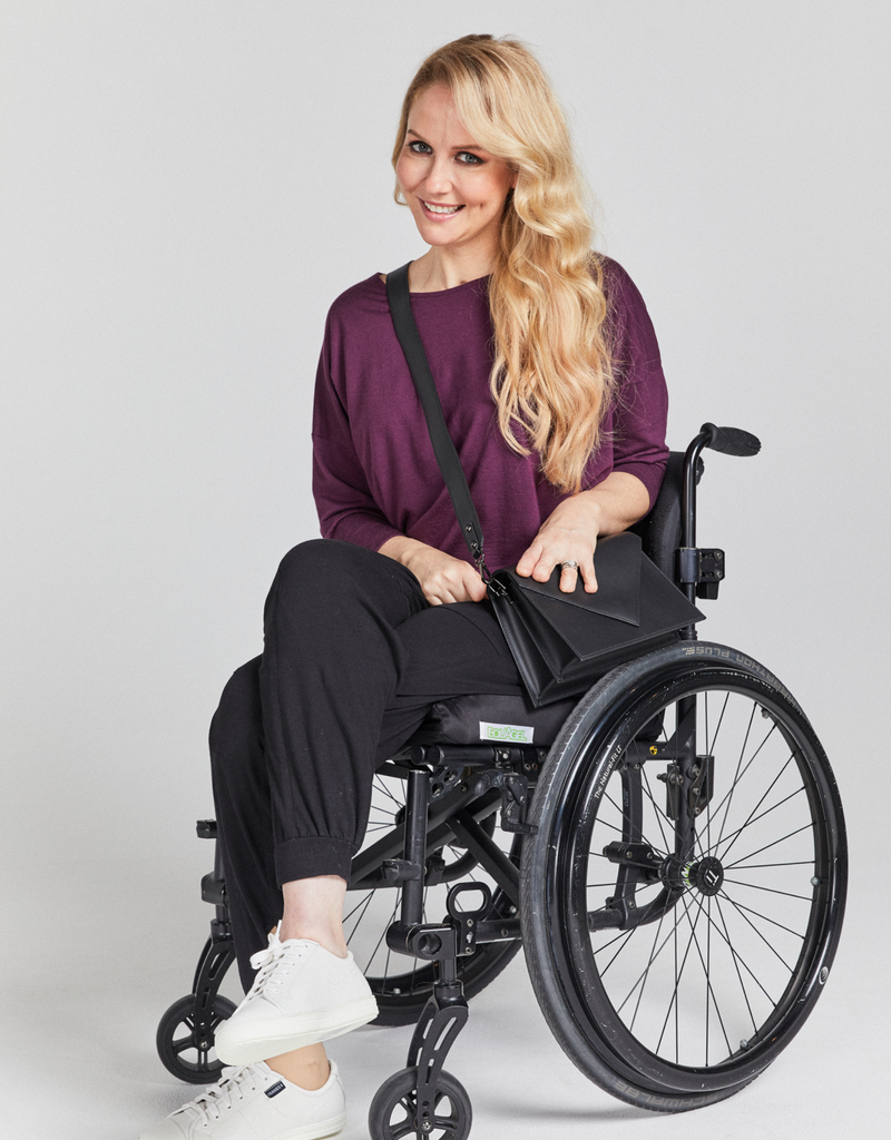 Blonde woman in wheelchair wearing a grape (purple) coloured 3/4 sleeve loose top, black pants and with a black handbag crossed over her body.