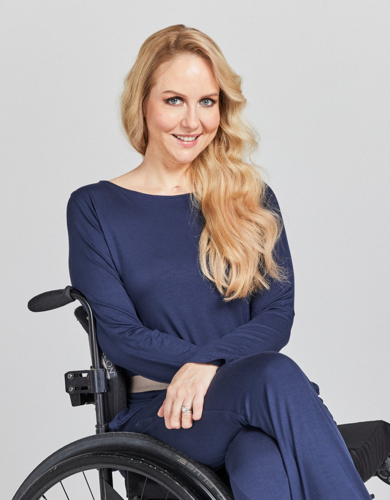 Blonde woman in wheelchair wearing a blue loose fitting, long sleeve top with rounded neckline. 