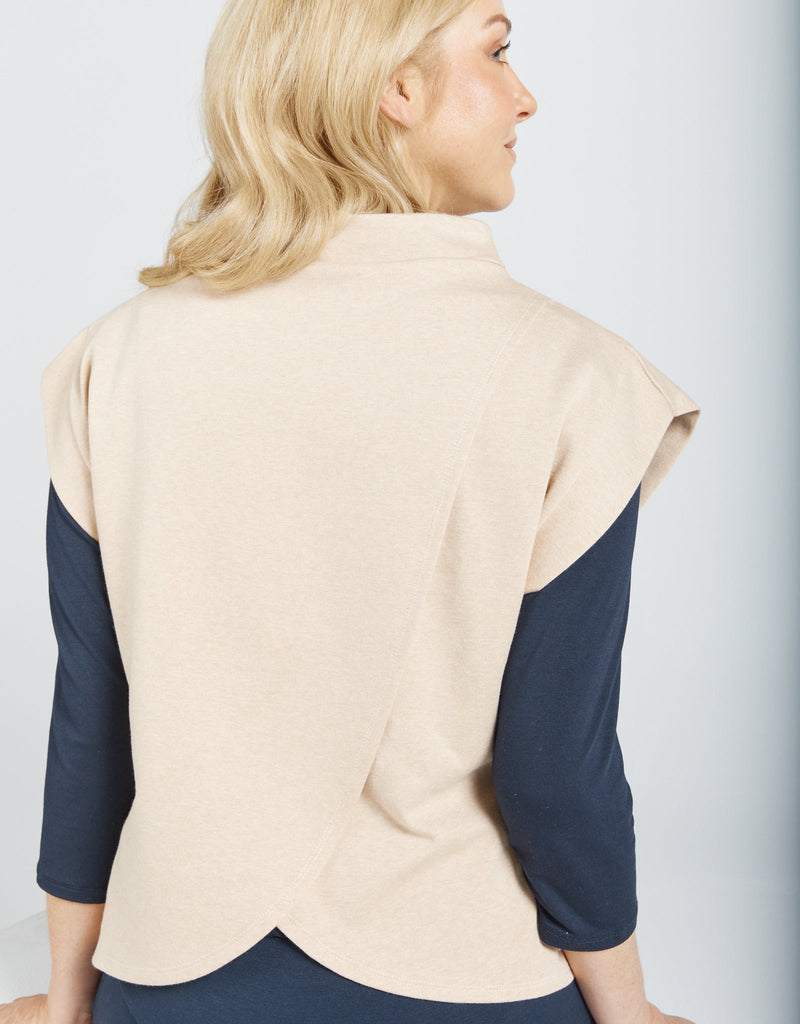 Image of a seated female with blonde hair wearing a natural cotton (beige colour) bat wing top with a high neckline. She has layered the top with a long sleeve navy blue shirt .Christina Stephens Adaptive Clothing Australia.