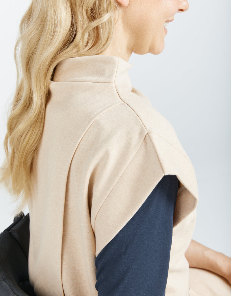 Image of a seated female in a wheelchair, with blonde hair wearing a natural cotton (beige colour) bat wing dress, with a high neckline. She has layered the top with a long sleeve navy blue shirt .Christina Stephens Adaptive Clothing Australia.