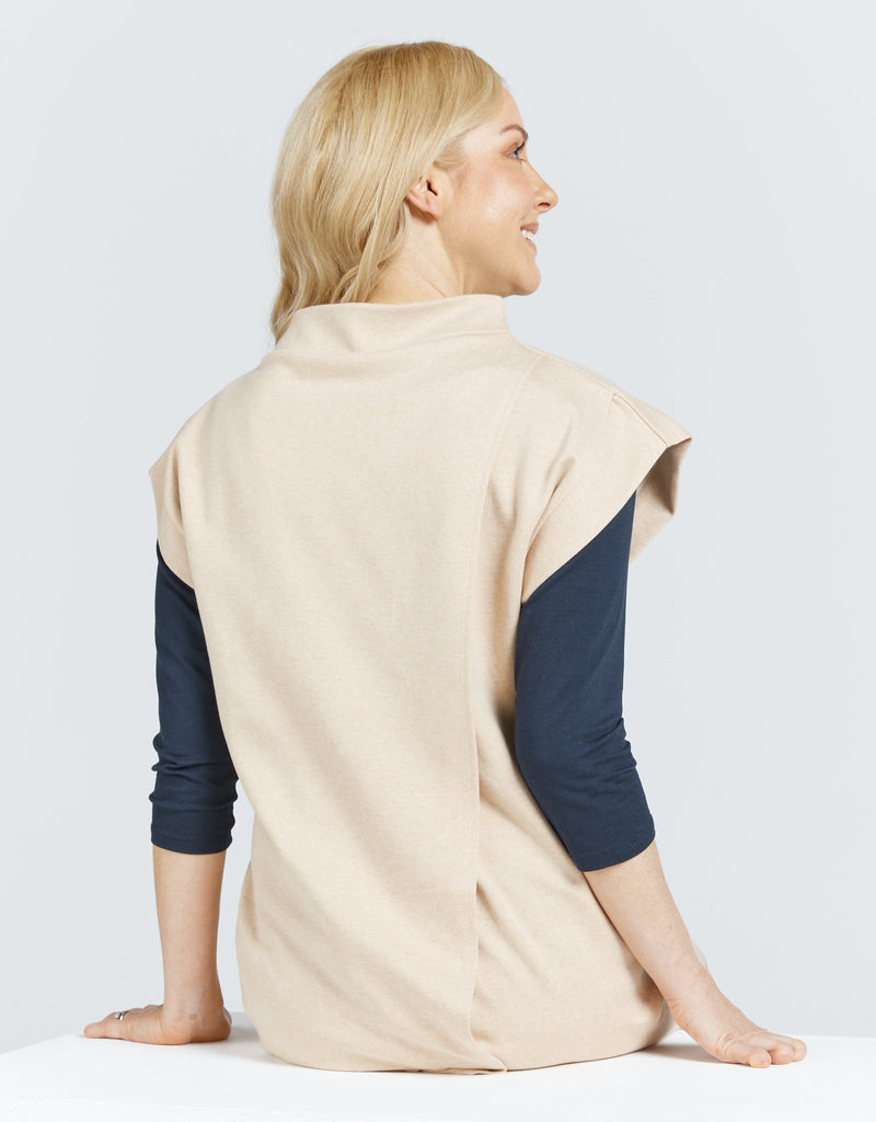 Image of a seated female in a wheelchair, with blonde hair wearing a natural cotton (beige colour) bat wing dress, with a high neckline. She has layered the top with a long sleeve navy blue shirt .Christina Stephens Adaptive Clothing Australia.