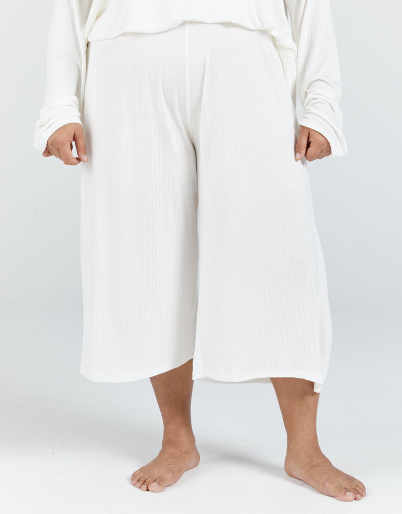 Image of a dark haired, dark skinned standing female, wearing a cream long sleeve top and cream cropped wide leg pants.Christina Stephens Adaptive Clothing Australia.