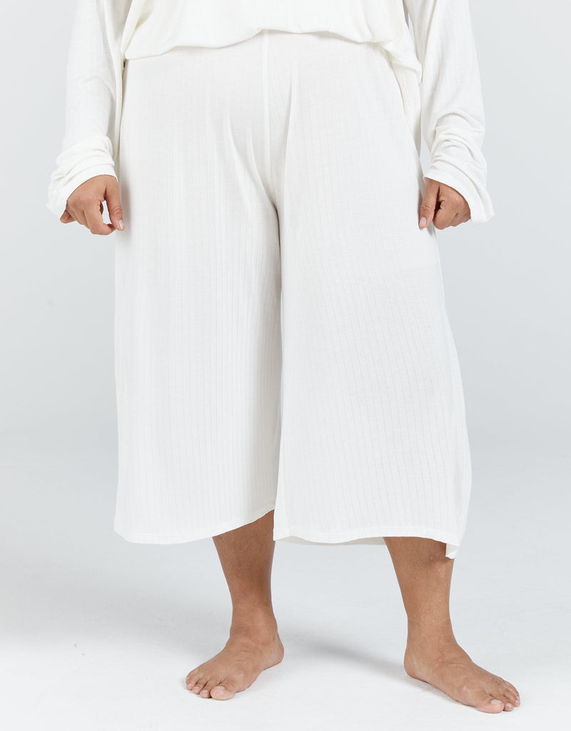 Image of a dark haired, dark skinned standing female, wearing a cream long sleeve top and cream cropped wide leg pants.Christina Stephens Adaptive Clothing Australia.