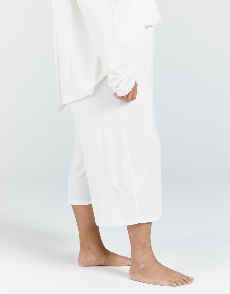 Image of a dark haired, dark skinned standing female, wearing a cream long sleeve top and cream cropped wide leg pants.Christina Stephens Adaptive Clothing Australia.