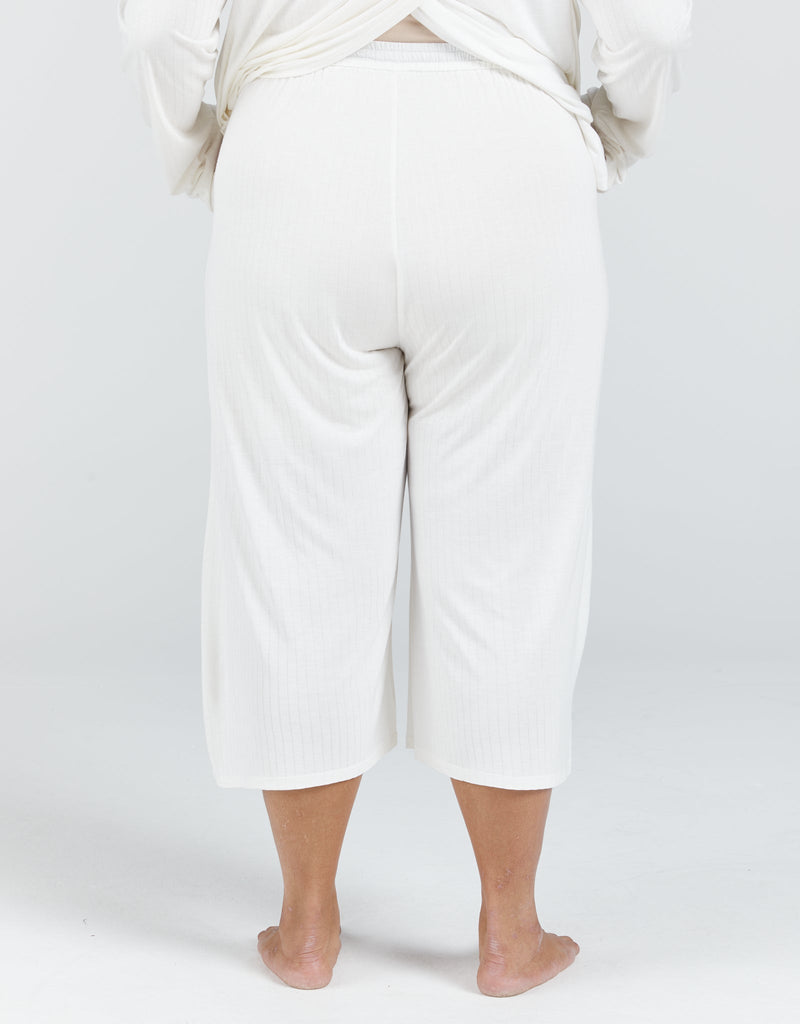 Image of a dark haired, dark skinned standing female, wearing a cream long sleeve top and cream cropped wide leg pants. She is facing backwards. Christina Stephens Adaptive Clothing Australia.