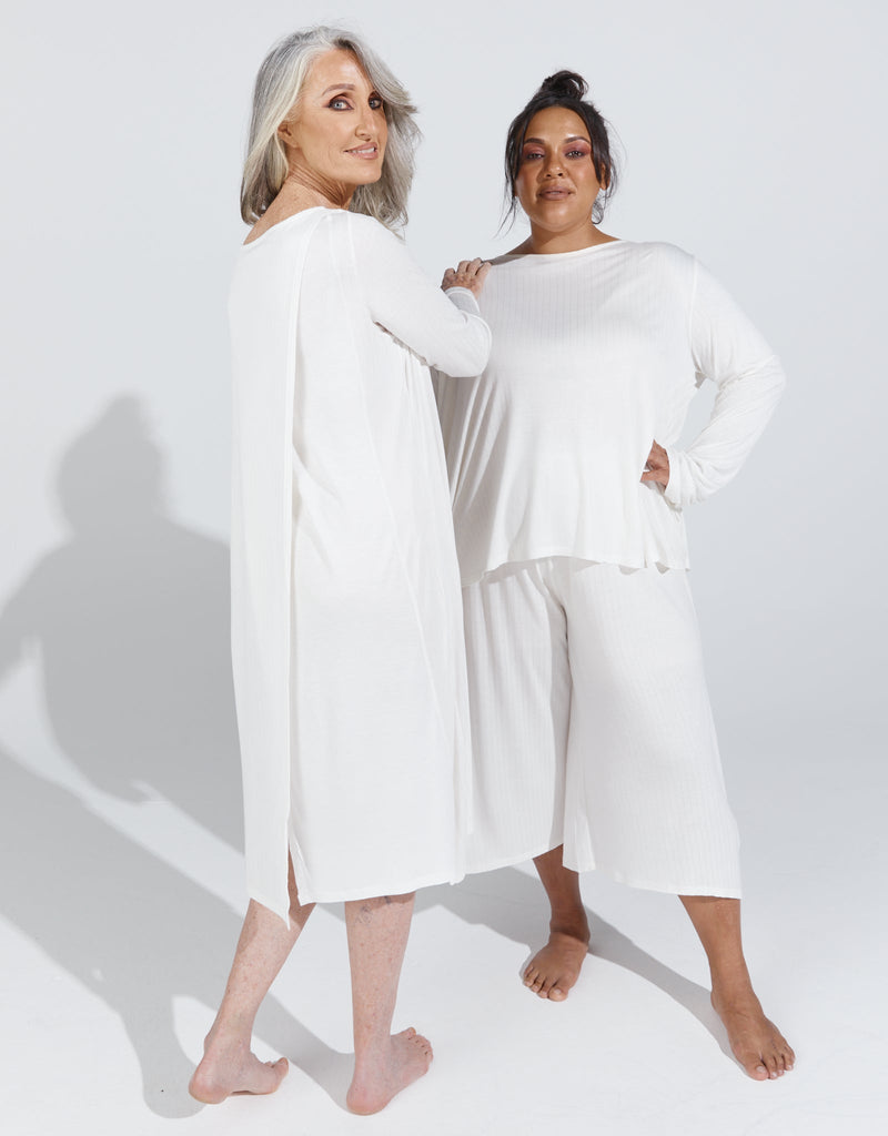 Image of a standing female with grey hair wearing a cream knee length, long sleeved night gown. She is standing next to a dark haired, dark skinned standing female, wearing a cream long sleeve top and cream cropped wide leg pants.Christina Stephens Adaptive Clothing Australia. 