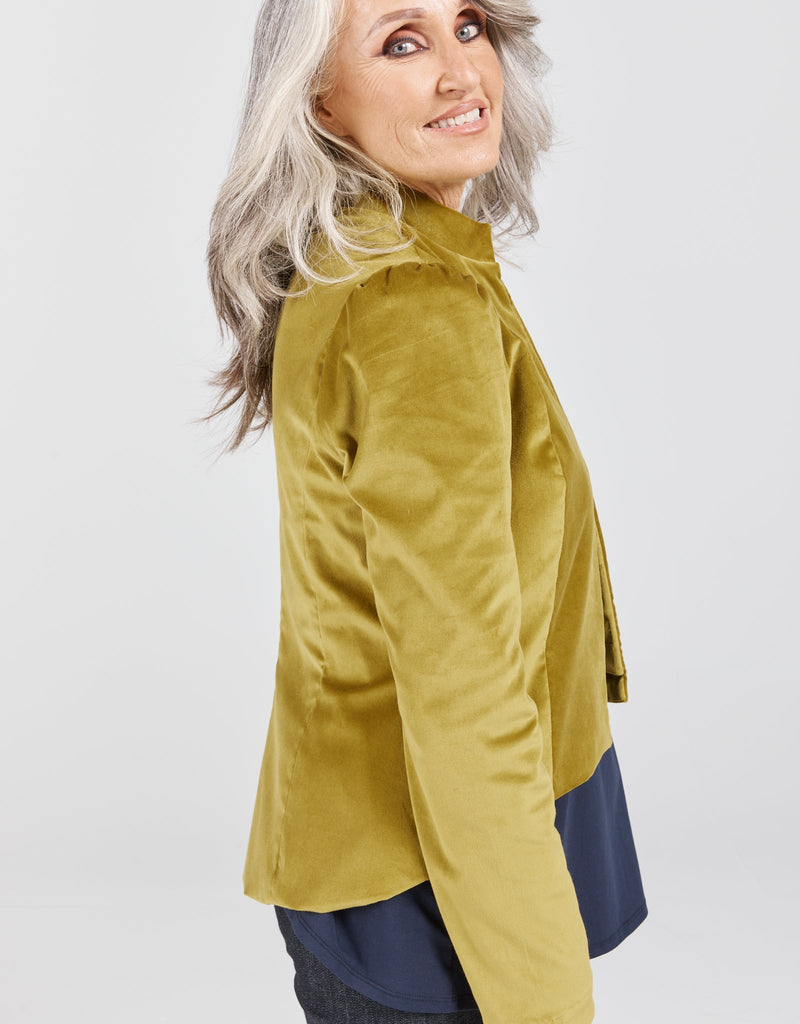 Image of a standing female with grey hair, wearing a citrus green velvet jacketing, a navy top and blue denim jeans. Christina Stephens Adaptive Clothing Australia. 