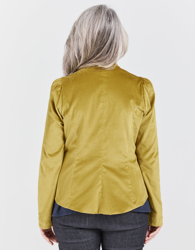 Image of a standing female with grey hair, wearing a citrus green velvet jacketing, a navy top and blue denim jeans.  She is facing backwards.Christina Stephens Adaptive Clothing Australia. 