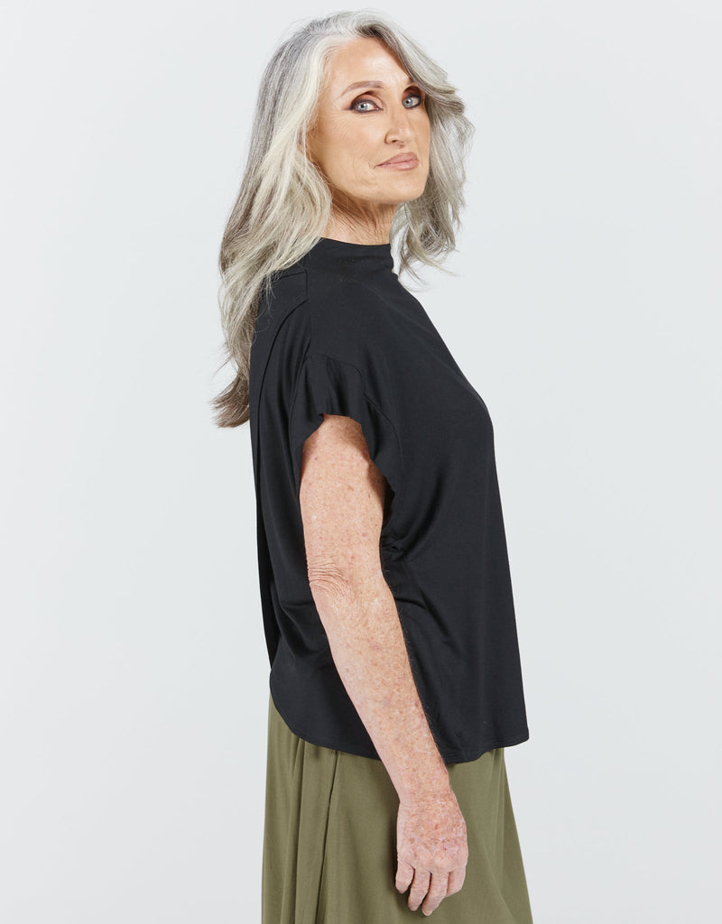 Image of a standing female with grey hair wearing a bat wing black high neck top with a khaki green skirt .Christina Stephens Adaptive Clothing Australia.