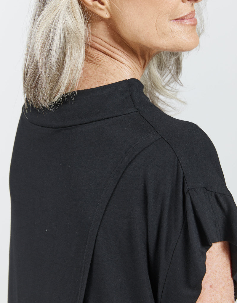 Image of a standing female with grey hair wearing a bat wing black high neck top. Christina Stephens Adaptive Clothing Australia.
