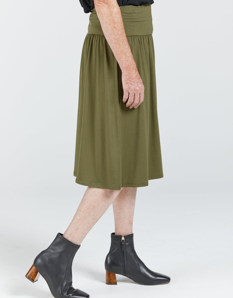 A standing lady is facing side-on and wearing a khaki bamboo skirt that reaches past her knees. It has a wide waist band and gathers below the band. She is wearing a black top and boots. Christina Stephens Adaptive Clothing Australia. 