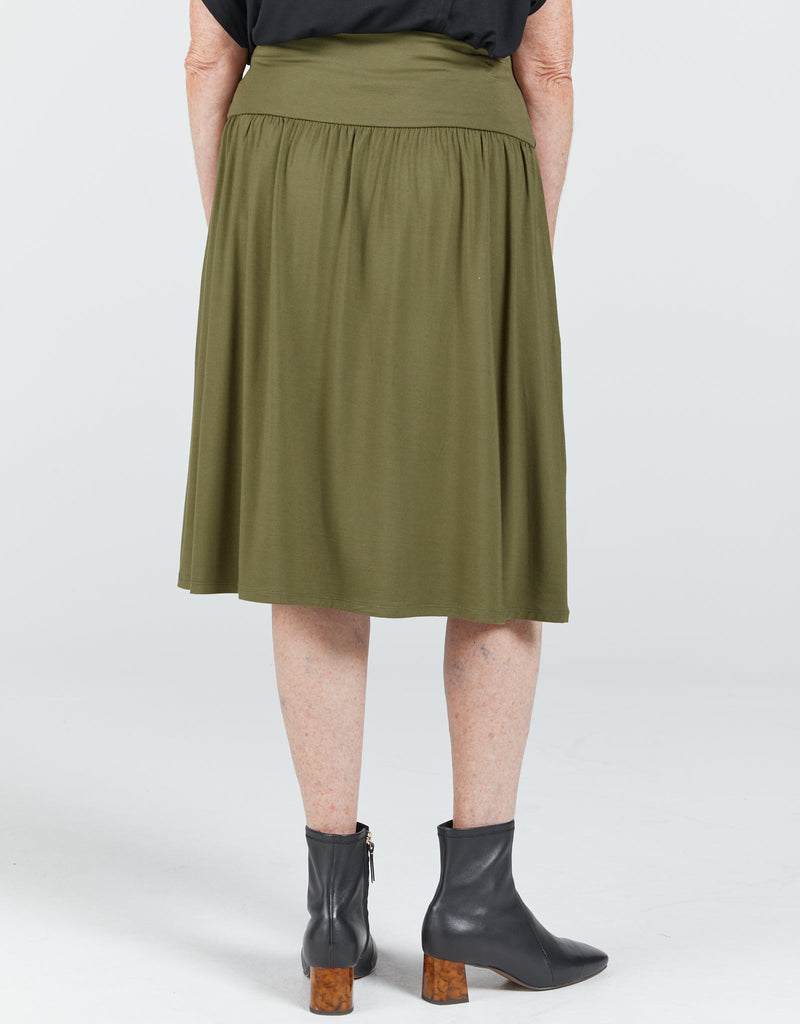 A standing lady facing towards the back is wearing a khaki bamboo skirt that reaches past her knees. It has a wide waist band and gathers below the band. She is wearing a black top and boots. Christina Stephens Adaptive Clothing Australia. 