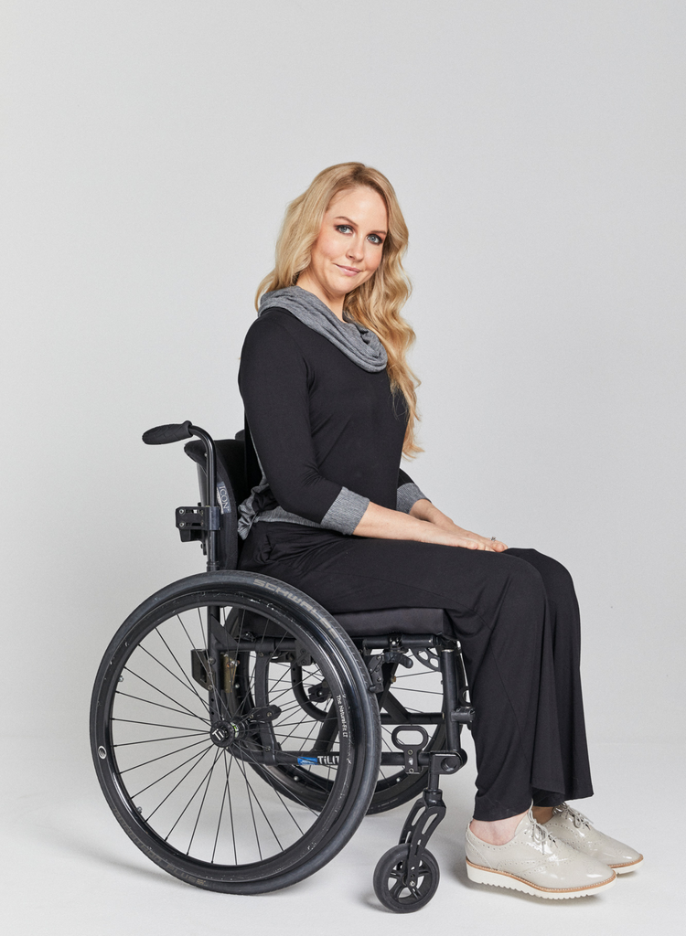Blonde woman in a wheelchair wearing black, bamboo leaf back top with grey stripe cowl neck and sleeve cuffs and black pants. Side facing. 