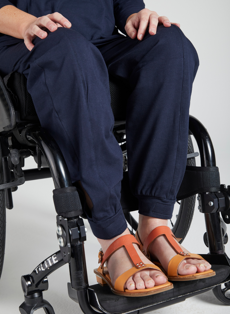 Model wearing navy pants and orange sandals and is seated in a wheelchair with her hands resting on her knees. Image is taken from waist down to the bottom of the wheelchair.
