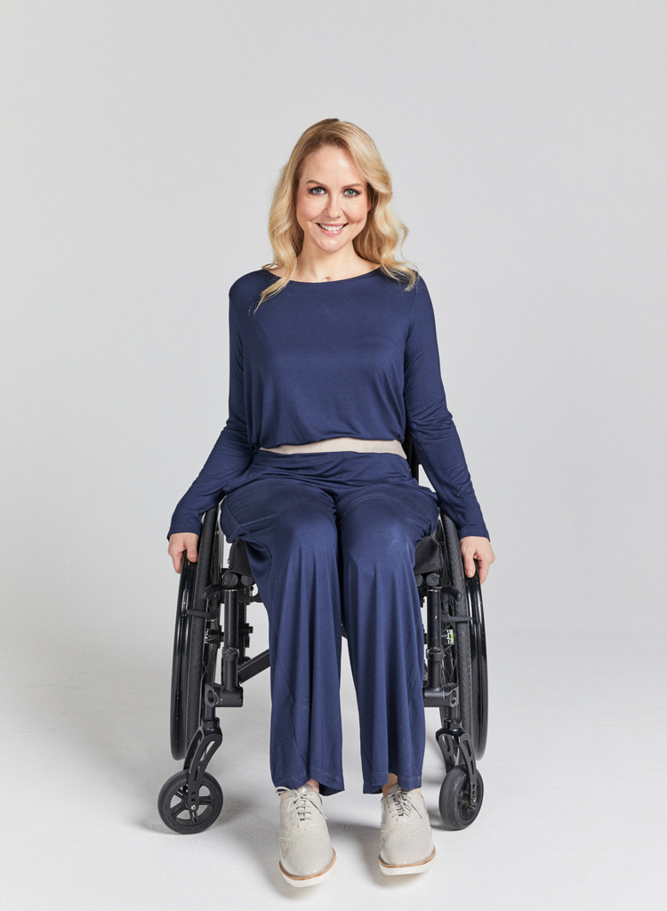 Blonde woman in a wheelchair wearing a blue and beige bamboo loose fitting top with long sleeves and blue loose fitting pants. Forward facing. Smiling.
