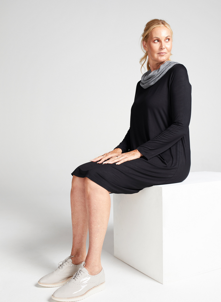 Blonde woman wearing a knee length, black long sleeve dress with grey stripe cowl neck. Sitting down on a white block, side facing. 