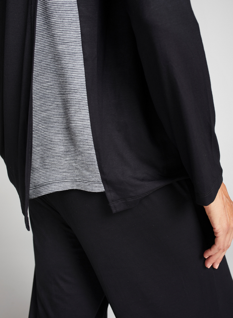 Brunette woman wearing a loose fitting black top with round neck line and long sleeves. Standing. Side facing. Side of garment detail (grey stripe underlay) in focus.
