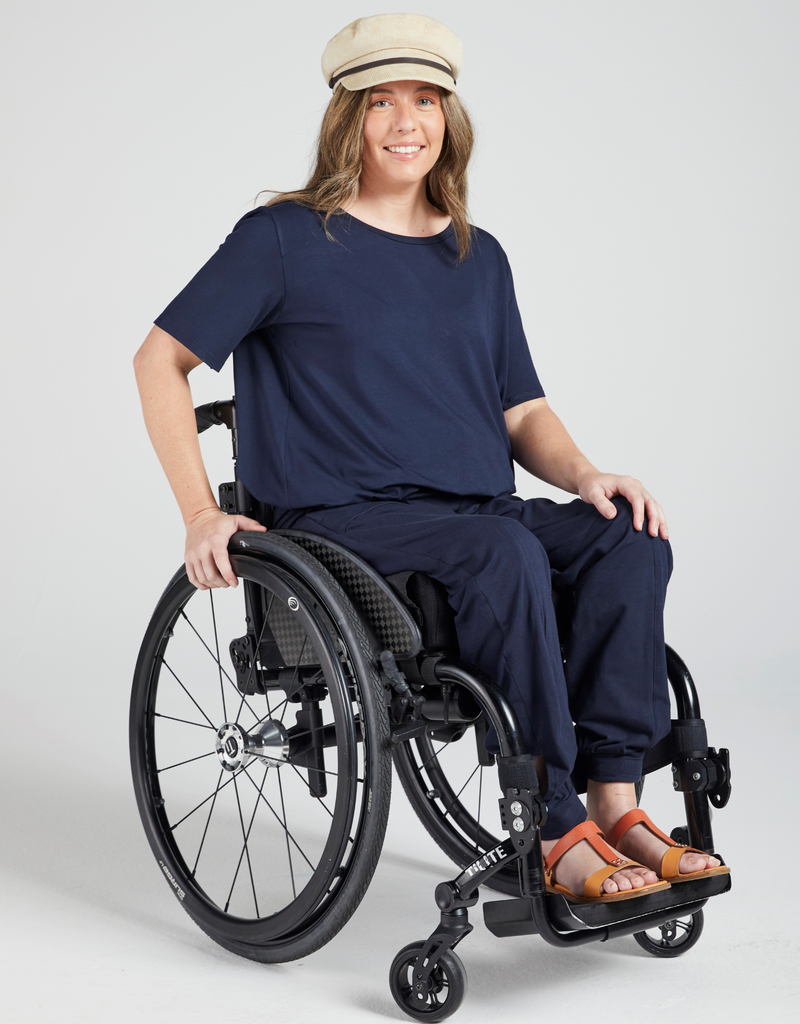 Dark blonde woman in a wheelchair wearing a navy, short sleeve t-shirt, beige cap and navy pants. Orange sandels on her feet. Forward facing and smiling. 
