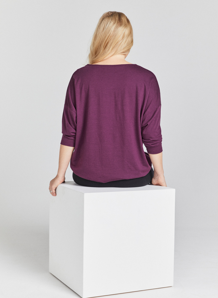 Seated blonde woman  wearing a grape (purple) round neck, loose top with 3/4 length sleeves and black pants. Back facing. 