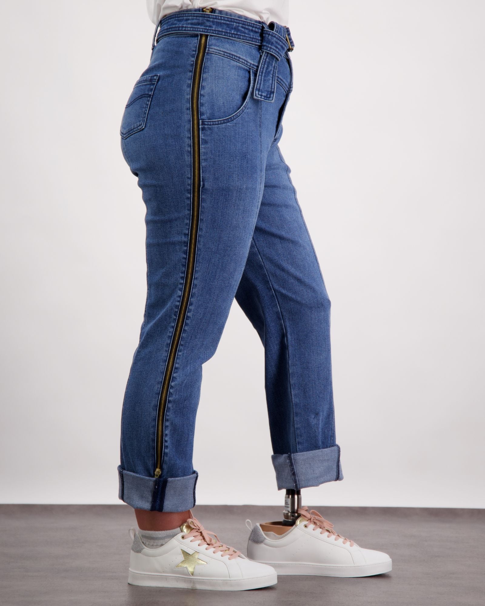 Christina Prosthetic Jeans featuring full-length side zippers, shown in medium wash denim with cuffed ankles. Adaptive design showcases innovative tear-resistant fabric and accessibility features, paired with white sneakers and a casual white top.