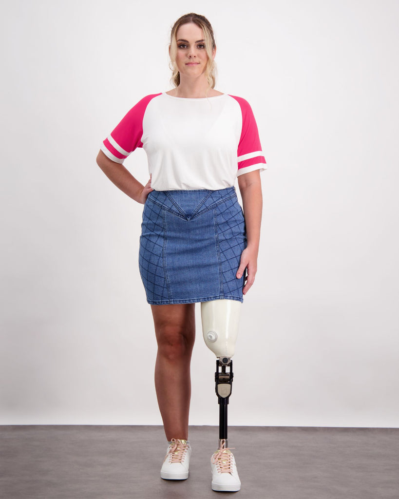 Female wearing a pink and white t-shirt and quilted blue denim skirt. She is wearing a prosthetic limb that is white and black. She is wearing white sneakers. Christina Stephens Adaptive Clothing Australia. 