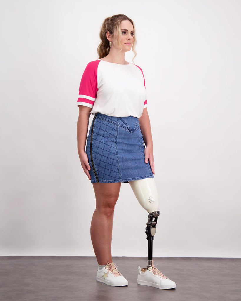 Female wearing a pink and white t-shirt and quilted blue denim skirt. She is wearing a prosthetic limb that is white and black. She is wearing white sneakers. Christina Stephens Adaptive Clothing Australia. 