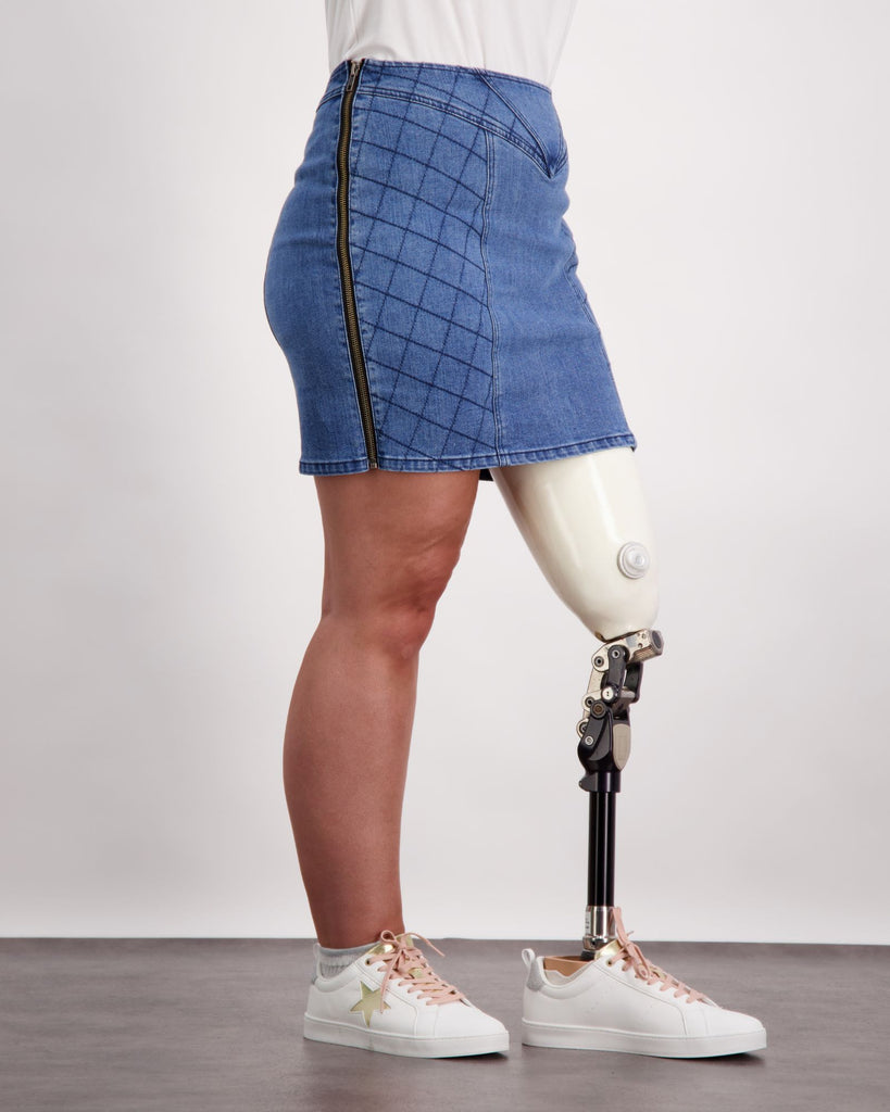 Female wearing a pink and white t-shirt and quilted blue denim skirt. She is wearing a prosthetic limb that is white and black. She is wearing white sneakers. Christina Stephens Adaptive Clothing Australia. 