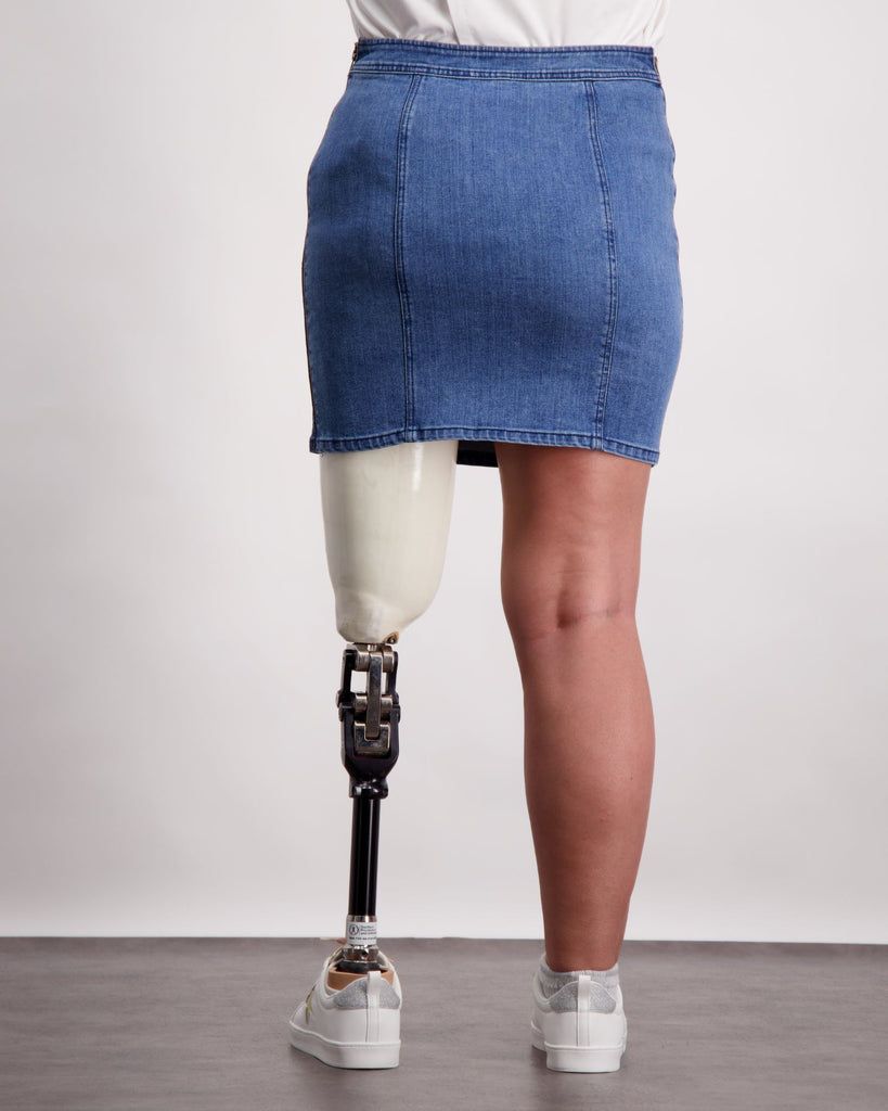 Female wearing a pink and white t-shirt and quilted blue denim skirt. She is wearing a prosthetic limb that is white and black. She is wearing white sneakers. Christina Stephens Adaptive Clothing Australia. 