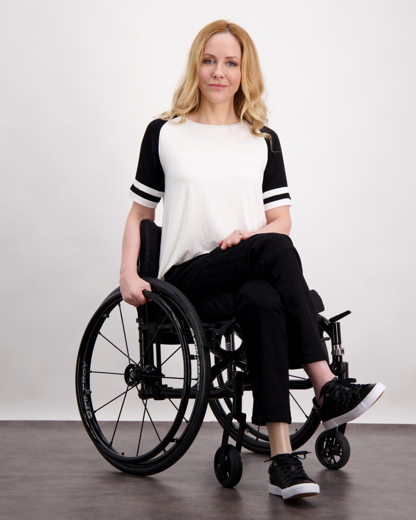 A blonde female seated in a wheelchair is wearing a white and black raglan t-shirt and a pair of black denim adaptive jeans with quilting on the knees and zips on the side of the legs. Full length image. Christina Stephens Australian Adaptive Clothing.  