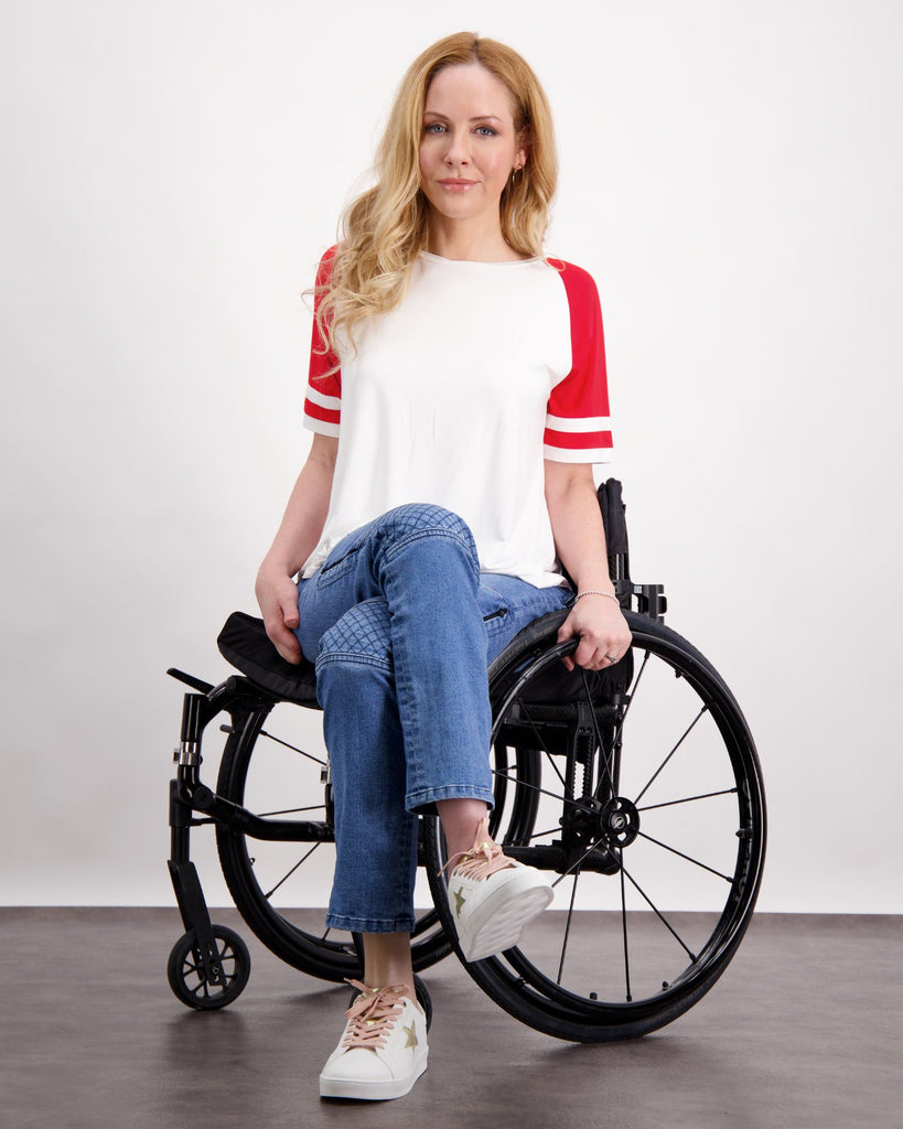 A blonde female seated in a wheelchair is wearing a white and red raglan t-shirt and a pair of blue denim adaptive jeans with quilting on the knees and zips on the side of the legs. Christina Stephens Australian Adaptive Clothing.  