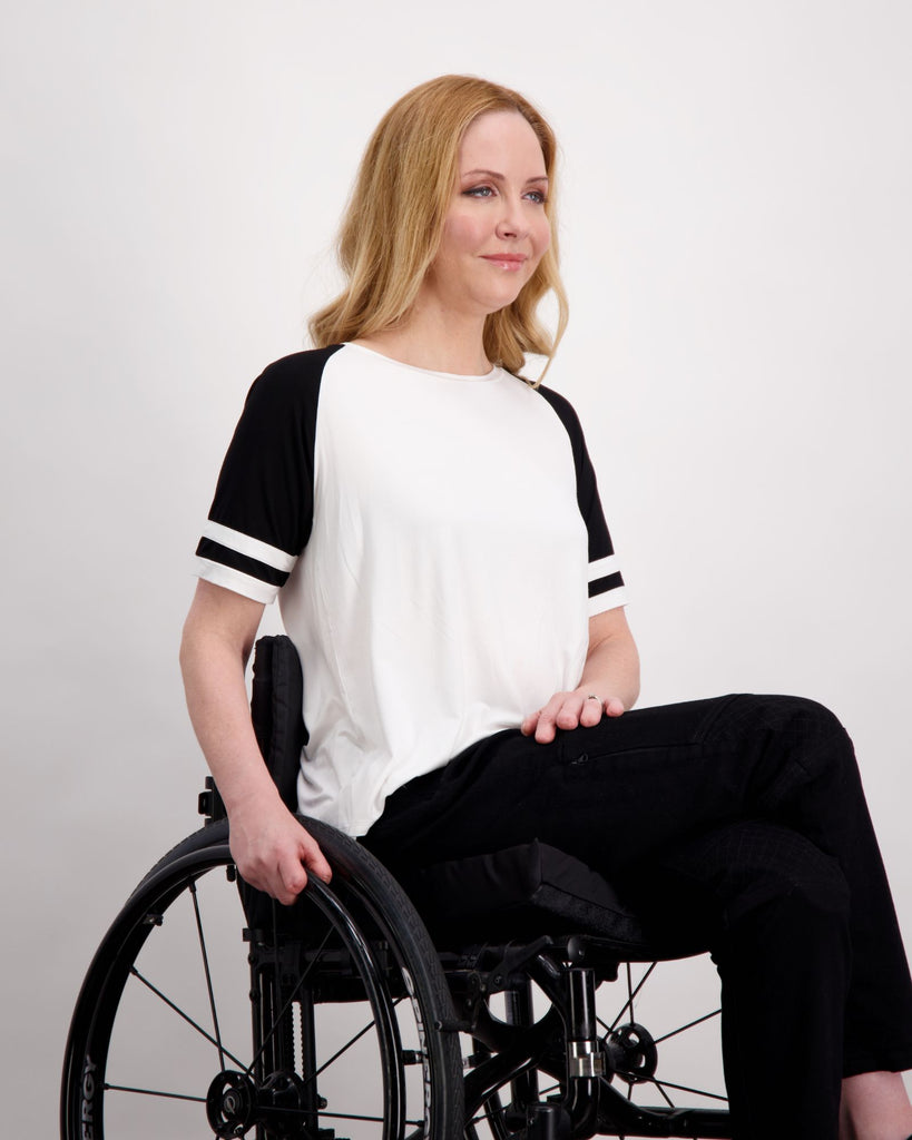 A blonde female seated in a wheelchair is wearing a white and black raglan t-shirt and a pair of black denim adaptive jeans with quilting on the knees and zips on the side of the legs. Christina Stephens Australian Adaptive Clothing.  