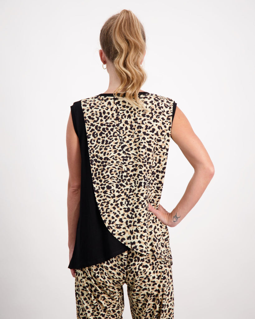 A standing blonde female wearing a pony-tail is wearing a black and leopard print bamboo sleeveless physio top.  She is facing towards the back of the room to highlight the leaf back design of the top. The back of the top has an upper leaf that is leopard print and the bottom leaf is black fabric. She has paired the top with matching leopard print bamboo pants. Christina Stephens Australian Adaptive Clothing. 
