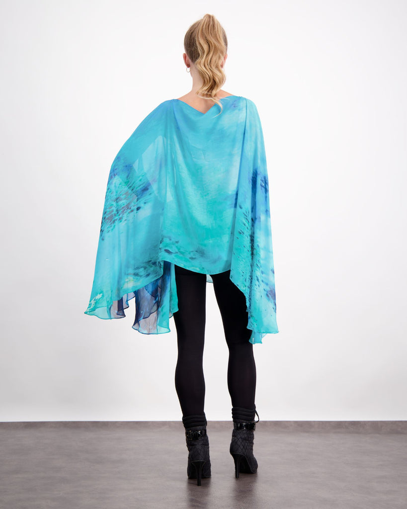 Blonde standing female is wearing a silk, aqua blue print kaftan with small pearl buttons, black leggings and black boots. She is facing to the back. Christina Stephens Adaptive Clothing Australia. 