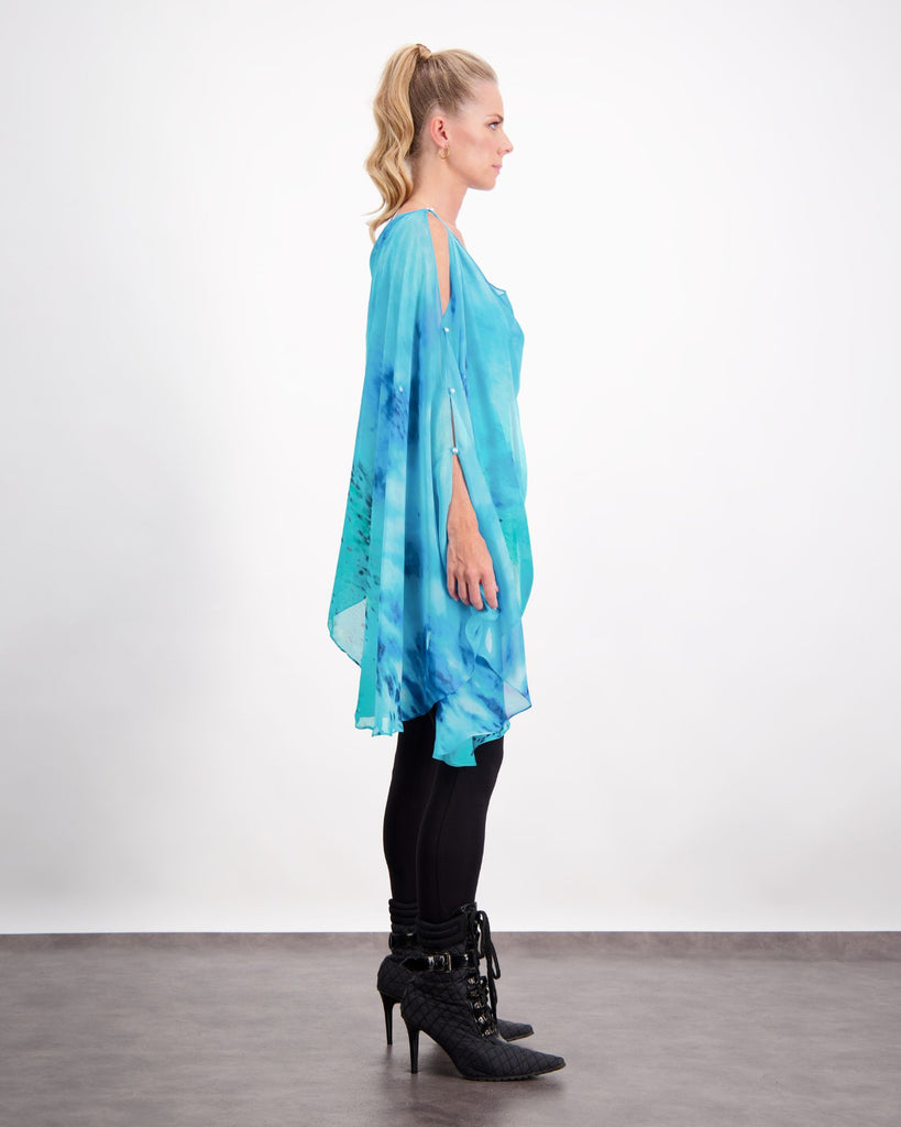 Blonde standing female is wearing a silk, aqua blue print kaftan with small pearl buttons, black leggings and black boots. Christina Stephens Adaptive Clothing Australia. 