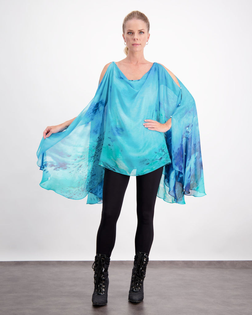 Blonde standing female is wearing a silk, aqua blue print kaftan with small pearl buttons, black leggings and black boots. Christina Stephens Adaptive Clothing Australia. 