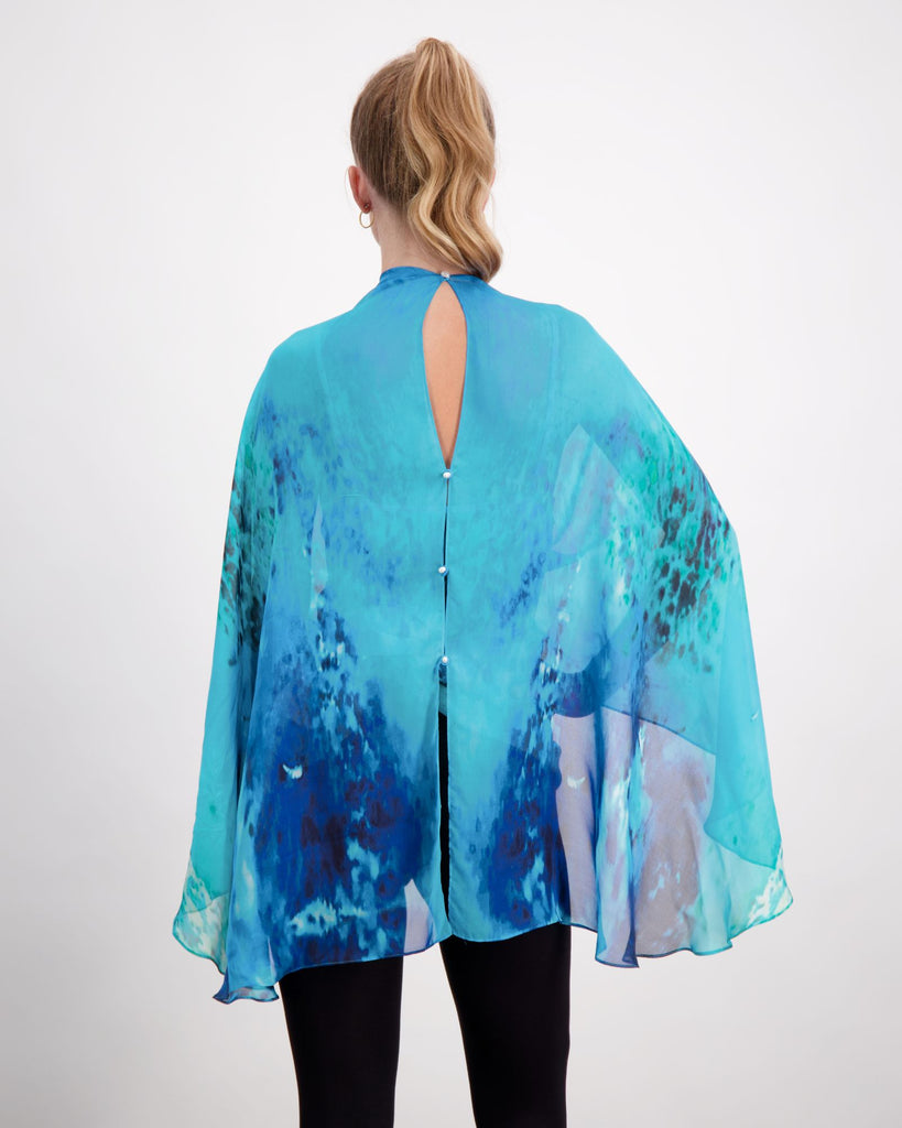 Blonde standing female is wearing a silk, aqua blue print kaftan with small pearl buttons, black leggings and black boots. Christina Stephens Adaptive Clothing Australia. 