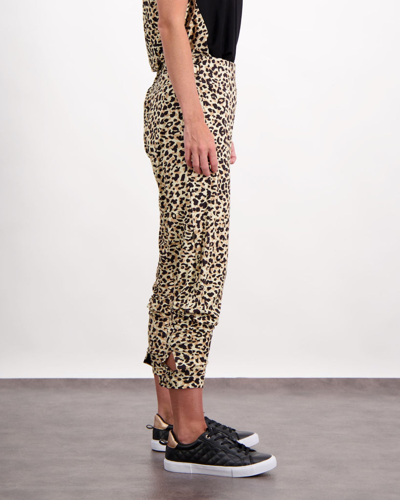 A standing woman is wearing leopard print physio leggings with a cuff at the bottom of the leg and wide pull tabs at her waist (in self-fabric). She is facing side-on. Styled so the leggings are pulled up to finish at her bottom of her shin. She is wearing black leather running shoes. The bottom of her black and leopard print top is visible. Christina Stephens Australian Adaptive Clothing. 