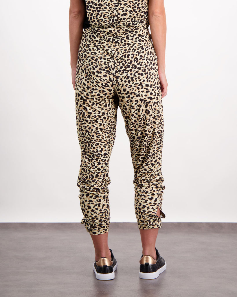 A standing woman is wearing leopard print physio leggings with a cuff at the bottom of the leg and a wide waist-band. She is facing towards the back of the room. The back of the garment has a thin, flat seam down the back. Styled so the leggings are pulled up to finish at her bottom of her shin. She is wearing black land gold leather running shoes. The bottom of her black and leopard print top is visible. Christina Stephens Australian Adaptive Clothing. 