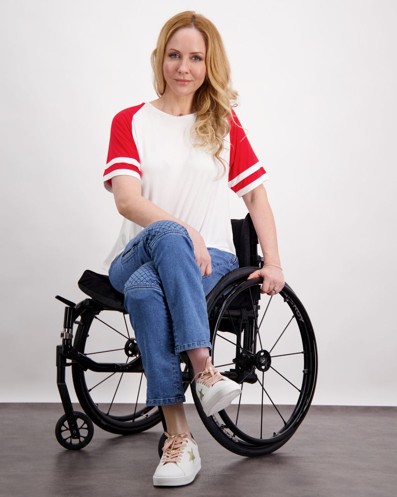 A blonde female seated in a wheelchair is wearing a white and red raglan t-shirt and a pair of blue denim adaptive jeans with quilting on the knees and zips on the side of the legs. Christina Stephens Australian Adaptive Clothing.  