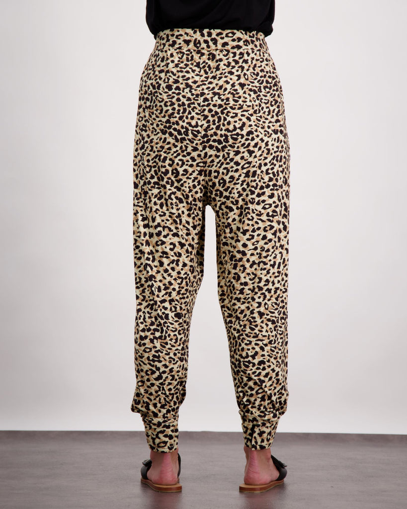 An image of a standing female wearing leopard print slouch pants black leather slippers. She is facing to the back of the room and you can see the backside of the slouch pant with a flat seam and wide waistband. The bottom of her black top is visible. Chistina Stephens Adaptive Clothing Australia. 