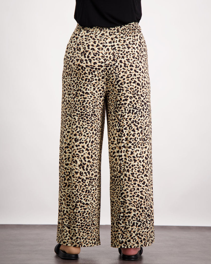 Image is of a female wearing a pair of leopard print bamboo wide leg pants and black leather slippers. She is facing towards the back of the room. The back elastic gather is visible on the pants. The bottom of her black top is visible. Christina Stephens Australian Adaptive Clothing.
