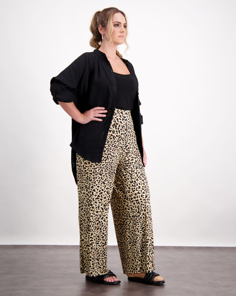 A standing female with caucasian skin and sandly coloured curly hair is wearing a black camisole under a black open linen shirt. She is wearing leopard print wide leg pants and black leather slippers. Christina Stephens Australian Adaptive Clothing.